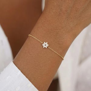 May include: A delicate gold bracelet featuring a star-shaped charm. The charm is encrusted with small, sparkling stones. The bracelet is worn on a wrist, and the background is a white shirt.