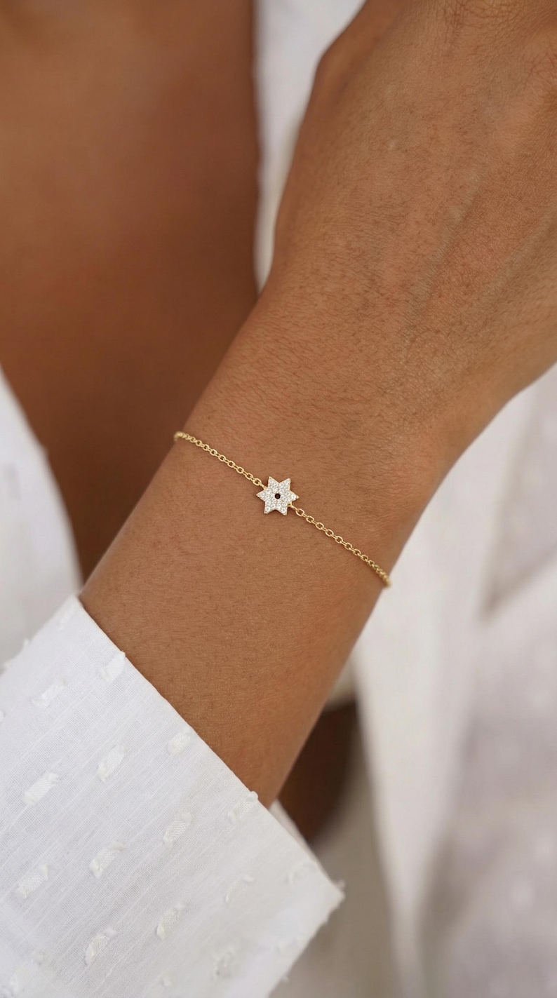 May include: A delicate gold bracelet featuring a star-shaped charm. The charm is encrusted with small, sparkling stones. The bracelet is worn on a wrist, and the background is a white shirt.