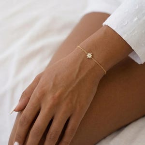 May include: A delicate gold bracelet featuring a star-shaped charm adorned with small, sparkling stones. The bracelet is worn on a wrist, showcasing its fine chain and elegant design. The jewelry piece adds a touch of sophistication.