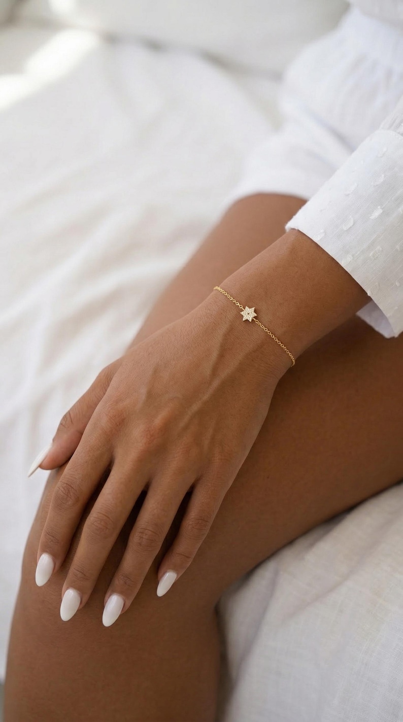 May include: A delicate gold bracelet featuring a star-shaped charm adorned with small, sparkling stones. The bracelet is worn on a wrist, showcasing its fine chain and elegant design. The jewelry piece adds a touch of sophistication.