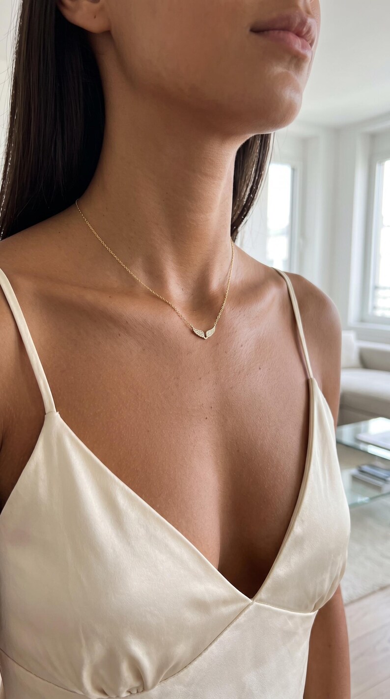 May include: A delicate gold necklace with a wing-shaped pendant, adorned with small, sparkling stones. The necklace is worn on a person's neck, against a cream-colored top.