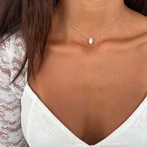 May include: A delicate silver necklace with a small, oval-shaped pendant. The pendant is centered on a fine chain, creating a minimalist and elegant look. The necklace is shown against a backdrop of a white lace top.