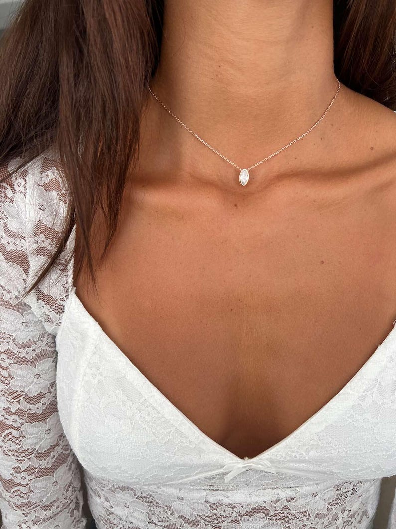 May include: A delicate silver necklace with a small, oval-shaped pendant. The pendant is centered on a fine chain, creating a minimalist and elegant look. The necklace is shown against a backdrop of a white lace top.