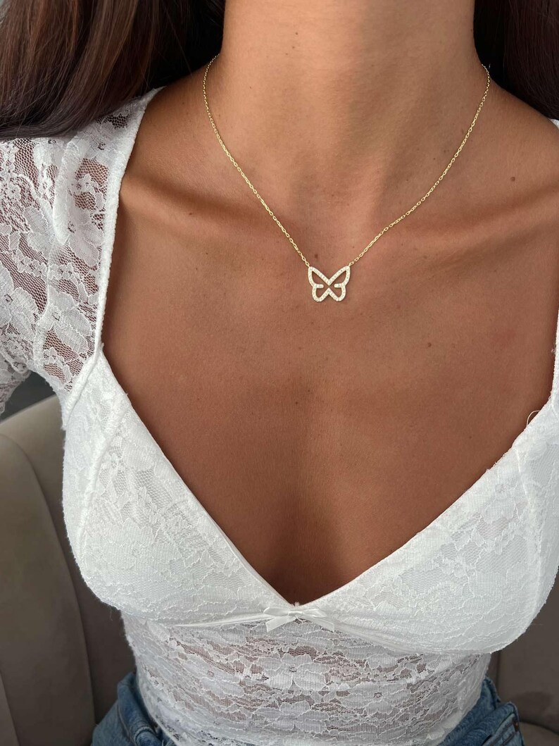 May include: A delicate gold-colored butterfly pendant necklace. The butterfly design is encrusted with small, sparkling stones. The necklace is displayed on a person's chest, paired with a white lace top.