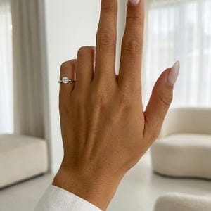 May include: A silver-toned ring featuring a single round-cut clear gemstone set in a prong setting. The ring is worn on the ring finger, showcasing a polished metal band that encircles the finger, highlighting the clarity and brilliance of the stone.