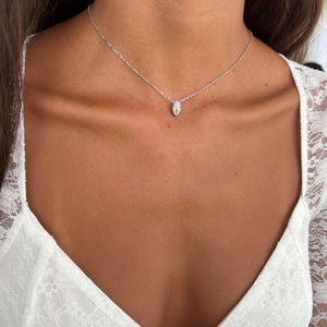 May include: A delicate silver necklace with a small, oval-shaped pendant. The pendant is centered on a fine chain, creating a minimalist and elegant look. The necklace is worn against a backdrop of a white lace top.