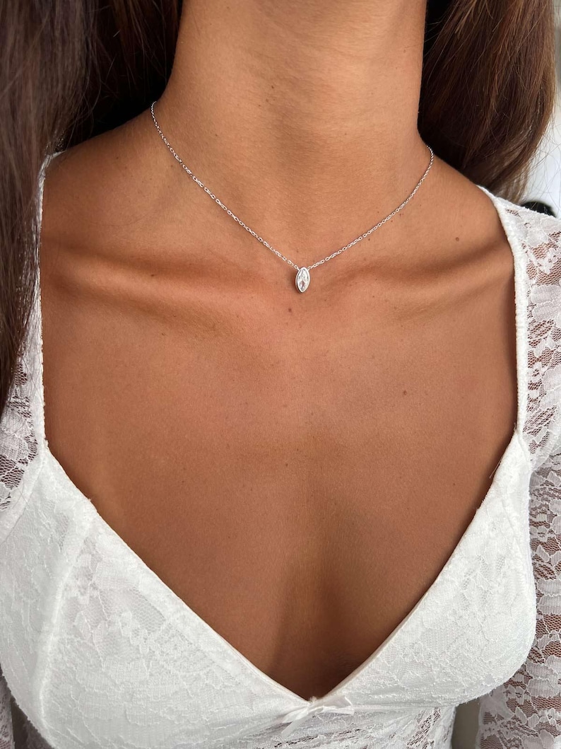 May include: A delicate silver necklace with a small, oval-shaped pendant. The pendant is centered on a fine chain, creating a minimalist and elegant look. The necklace is worn against a backdrop of a white lace top.