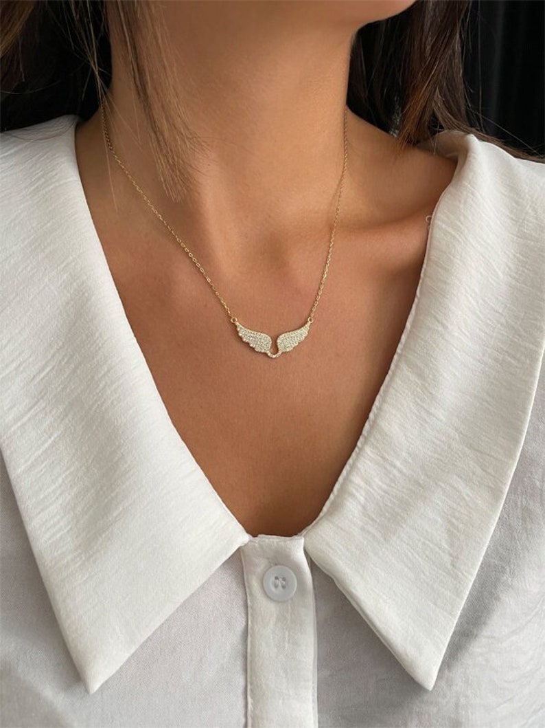 Puede incluir: Un collar de oro con un delicado colgante de ala brillante. El collar est&aacute; en una modelo que lleva una camisa blanca con cuello.