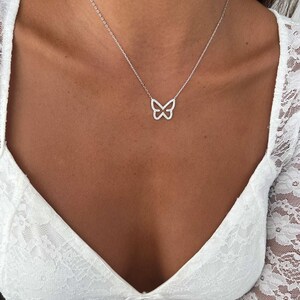 May include: A delicate silver necklace featuring a butterfly pendant. The butterfly design is outlined with small, sparkling stones. The necklace is worn against a white lace top, creating a simple yet elegant look. The chain is thin and subtle.