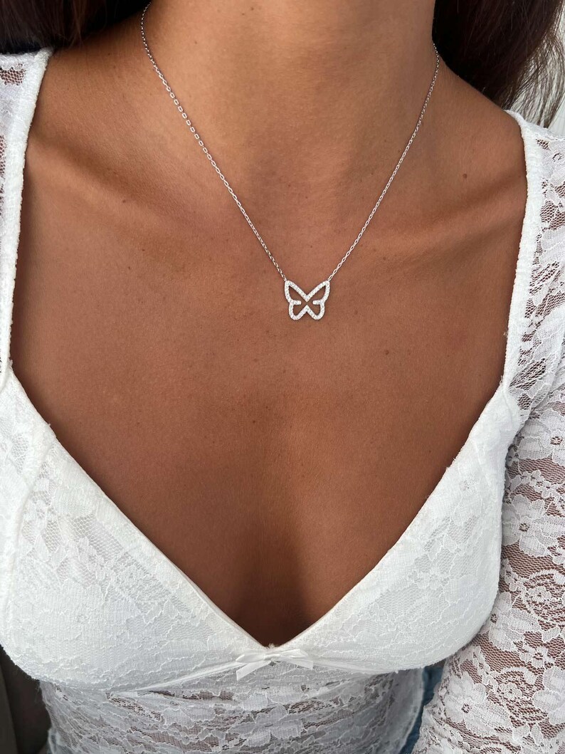 May include: A delicate silver necklace featuring a butterfly pendant. The butterfly design is outlined with small, sparkling stones. The necklace is worn against a white lace top, creating a simple yet elegant look. The chain is thin and subtle.