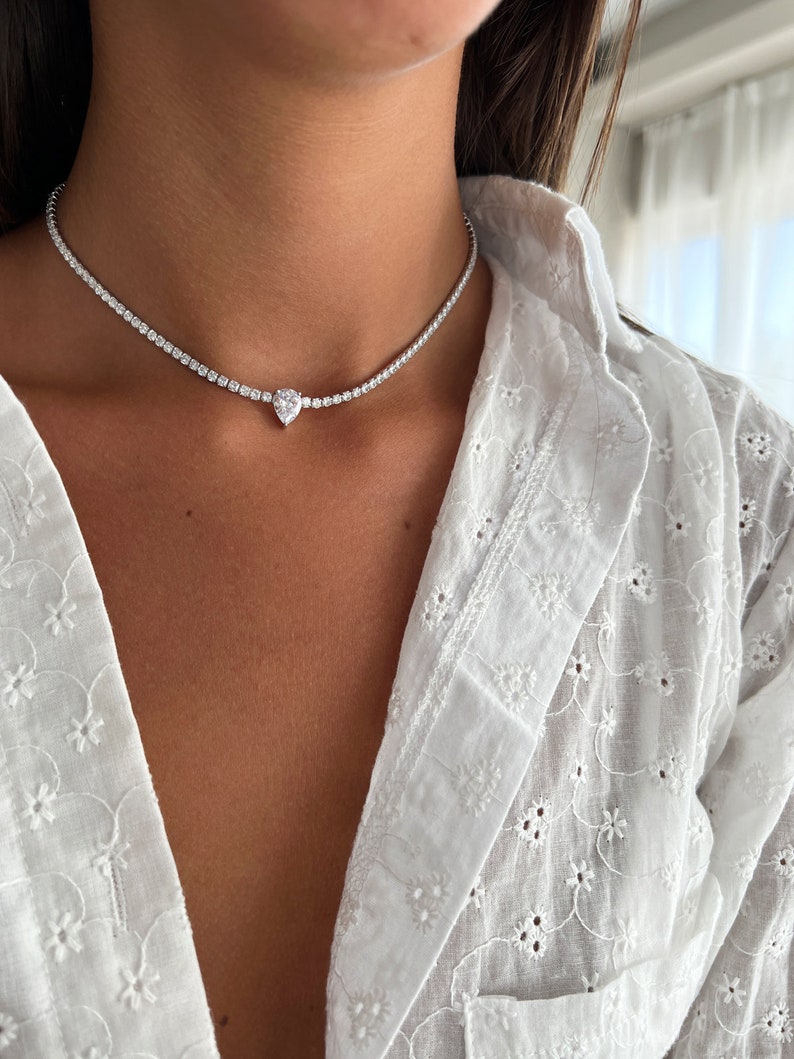Peut inclure: Un collier ras de cou argent&eacute; avec un pendentif en diamant de forme poire. Le collier est compos&eacute; de petites pierres claires.