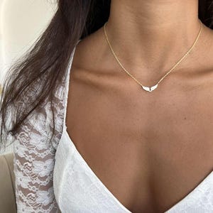 May include: A delicate gold necklace featuring a small, wing-shaped pendant. The necklace has a thin chain and is worn around the neck. The pendant is white and the chain is gold. The necklace is a piece of jewelry.