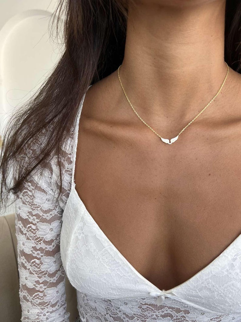 May include: A delicate gold necklace featuring a small, wing-shaped pendant. The necklace has a thin chain and is worn around the neck. The pendant is white and the chain is gold. The necklace is a piece of jewelry.