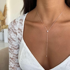 May include: A delicate rose gold necklace with a teardrop-shaped pendant and a dangling small gem. The necklace is worn over a white lace top, creating a simple yet elegant look. The necklace is a beautiful piece of jewelry.