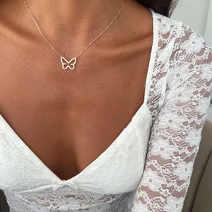 May include: A delicate gold-toned necklace featuring a butterfly pendant. The butterfly is outlined with small, sparkling stones. The necklace is worn against a backdrop of a white lace top.