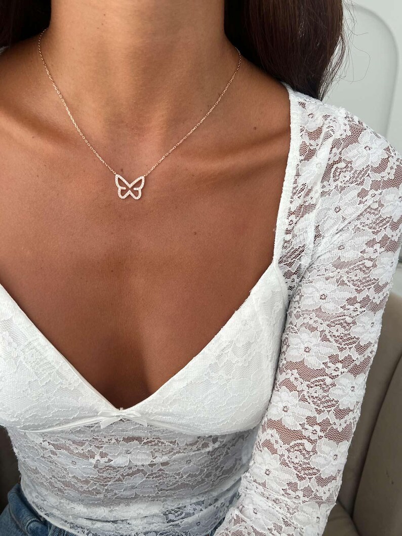 May include: A delicate gold-toned necklace featuring a butterfly pendant. The butterfly is outlined with small, sparkling stones. The necklace is worn against a backdrop of a white lace top.