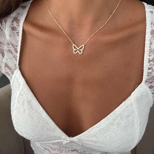 May include: A delicate gold-colored butterfly pendant necklace. The butterfly design is open, with a subtle sparkle. The necklace is worn on a person, against a white lace top.