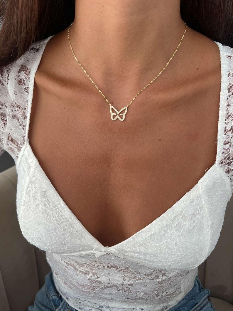 May include: A delicate gold-colored butterfly pendant necklace. The butterfly design is open, with a subtle sparkle. The necklace is worn on a person, against a white lace top.
