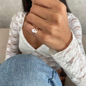 May include: Rose gold ring with a pear-cut, clear gemstone. The ring has a delicate double-band design. The ring is worn on a finger, highlighting its elegant and modern style. The gemstone is approximately 1 cm in length.