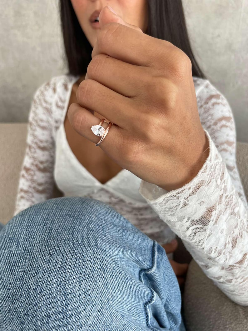 May include: Rose gold ring with a pear-cut, clear gemstone. The ring has a delicate double-band design. The ring is worn on a finger, highlighting its elegant and modern style. The gemstone is approximately 1 cm in length.