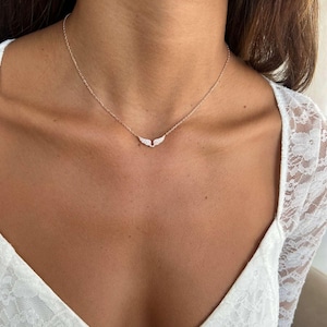 May include: A delicate rose gold necklace featuring a pendant shaped like small wings, adorned with sparkling clear crystals. The necklace is displayed against a white lace top, highlighting the jewelry's elegant design.