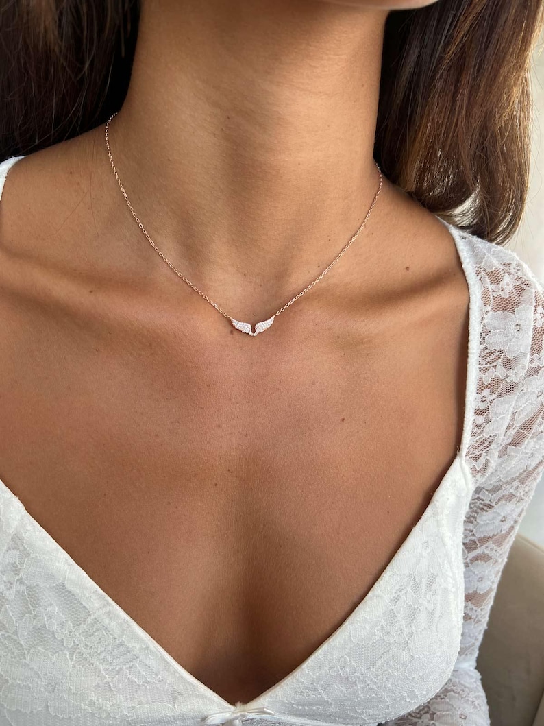 May include: A delicate rose gold necklace featuring a pendant shaped like small wings, adorned with sparkling clear crystals. The necklace is displayed against a white lace top, highlighting the jewelry's elegant design.
