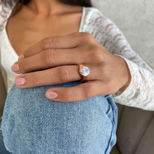 May include: A gold ring featuring a light blue, oval gemstone. The ring is worn on a finger. The background includes a white lace top and blue denim jeans.