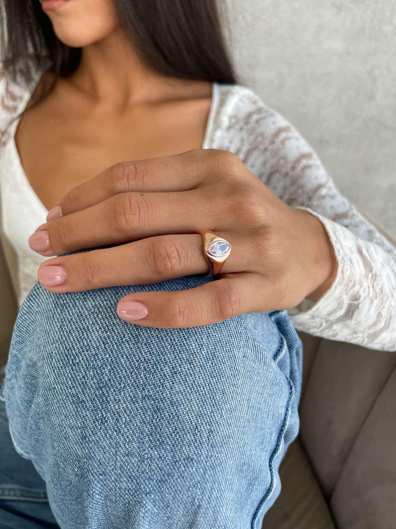May include: A gold ring featuring a light blue, oval gemstone. The ring is worn on a finger. The background includes a white lace top and blue denim jeans.