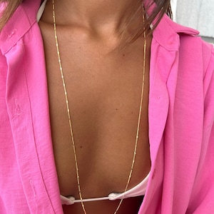 Könnte beinhalten: Eine lange, zarte Goldkette mit linearem Design. Die Halskette wird vor dem Hintergrund eines pinkfarbenen Hemdes mit Knöpfen und einem weißen Oberteil gezeigt.