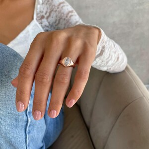 May include: A rose gold ring featuring a marquise-cut gemstone. The ring is worn on a hand with pink nail polish. The person is wearing a white lace top and blue jeans. The ring's design and rose gold colour are the main focus of the image.