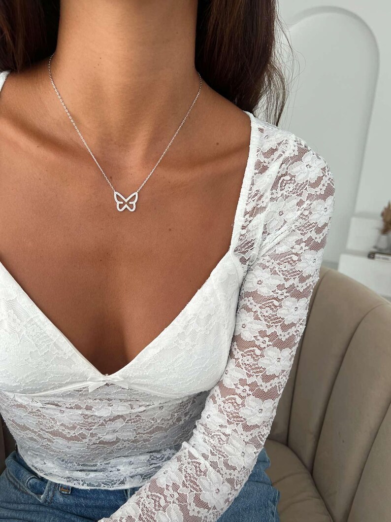 May include: A silver butterfly pendant necklace. The necklace features a delicate butterfly design suspended from a fine chain. The butterfly pendant is openwork, showcasing the intricate details of the design. The necklace is worn with a white lace top.