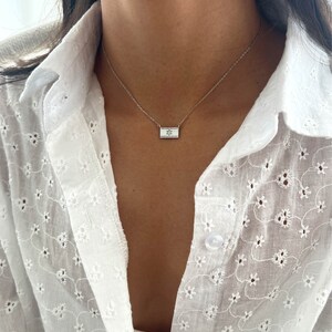 May include: A silver chain necklace with a small rectangular pendant featuring a star design. The necklace is worn by a person wearing a white, embroidered shirt.