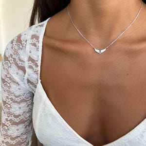 May include: A delicate silver necklace featuring a small pendant shaped like angel wings. The necklace is worn against a white lace top. The wings are detailed and the chain is thin.