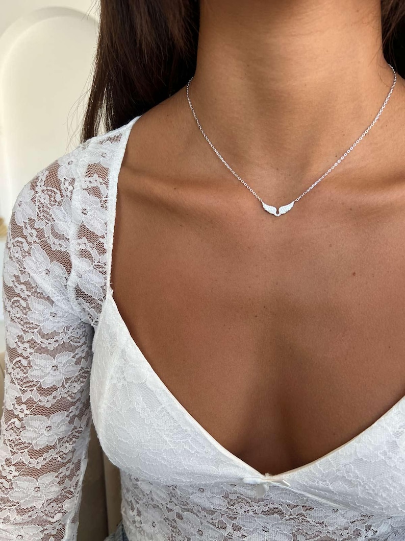 May include: A delicate silver necklace featuring a small pendant shaped like angel wings. The necklace is worn against a white lace top. The wings are detailed and the chain is thin.