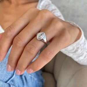 May include: A silver ring with a clear, marquise-shaped gemstone. The ring has a smooth, rounded band and a bezel setting. The ring is worn on a finger. The gemstone is set in a polished silver band.