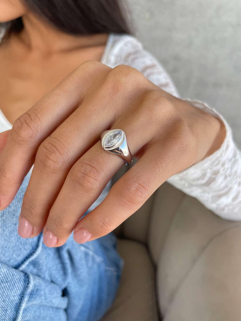 May include: A silver ring with a clear, marquise-shaped gemstone. The ring has a smooth, rounded band and a bezel setting. The ring is worn on a finger. The gemstone is set in a polished silver band.