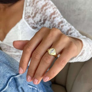 May include: A gold signet ring featuring a clear, oval-cut stone. The ring is worn on a finger with pink nail varnish. The background includes a white lace top and blue jeans.