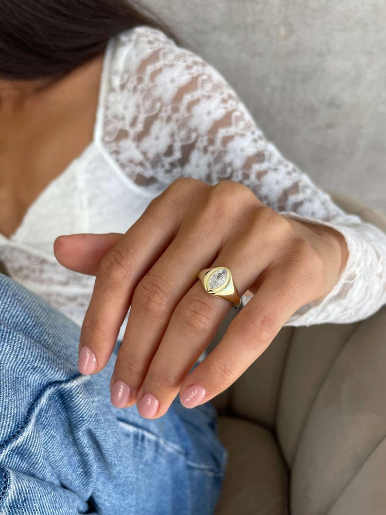 May include: A gold signet ring featuring a clear, oval-cut stone. The ring is worn on a finger with pink nail varnish. The background includes a white lace top and blue jeans.