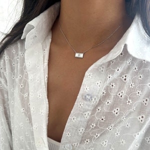 May include: A silver chain necklace with a small rectangular pendant featuring a star design. The necklace is worn by a person wearing a white, floral embroidered shirt.