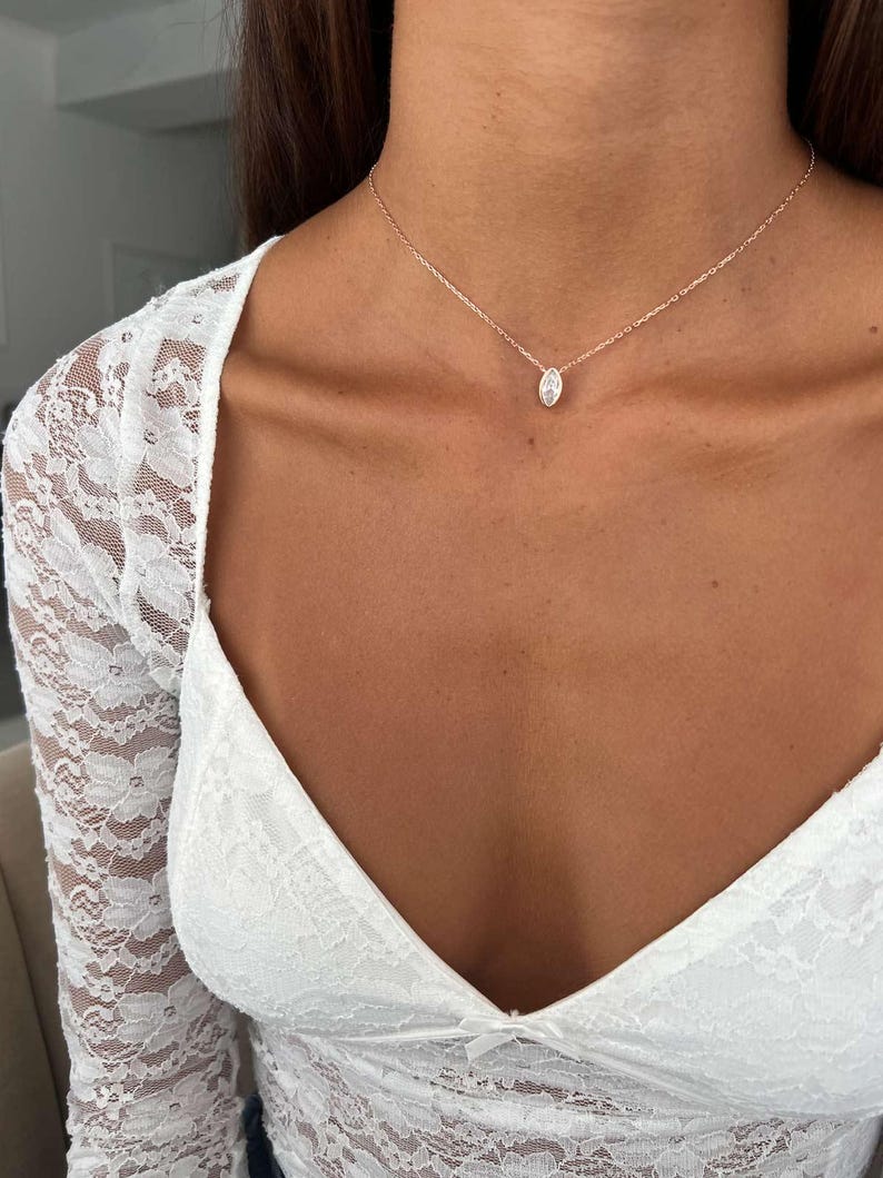 May include: A delicate rose gold necklace with a small, oval-shaped pendant. The pendant is centered on the neckline. The necklace is worn with a white lace top.