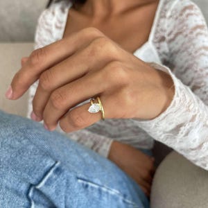 May include: A gold ring featuring a pear-cut, clear gemstone. The ring's band splits into two parts, one encircling the finger and the other supporting the gemstone. The ring is worn on a finger.