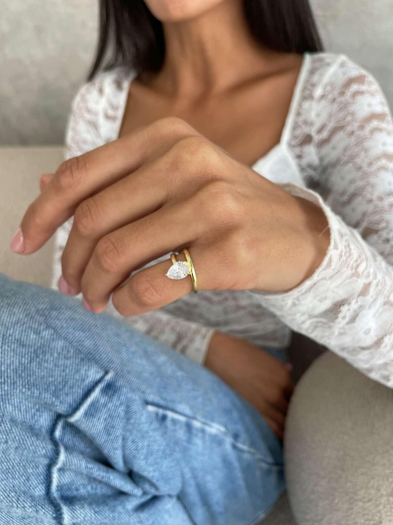 May include: A gold ring featuring a pear-cut, clear gemstone. The ring's band splits into two parts, one encircling the finger and the other supporting the gemstone. The ring is worn on a finger.