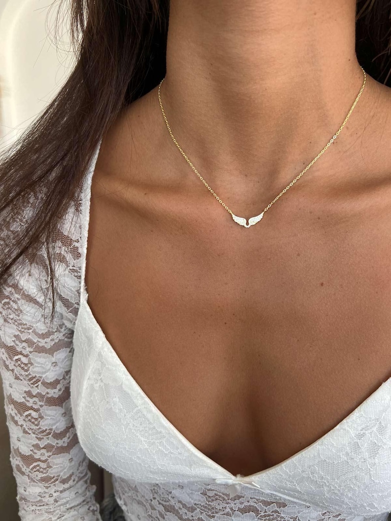 May include: A delicate gold-toned necklace featuring a pendant shaped like open wings, encrusted with small, sparkling stones. The necklace is worn against a backdrop of a white lace top, creating a contrast of textures and colors.