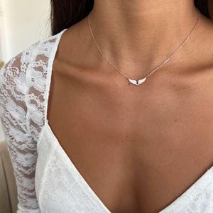 May include: A delicate necklace featuring a small, wing-shaped pendant. The pendant is adorned with small, sparkling stones. The necklace is gold-toned and rests on the neckline. The background includes a white lace top.