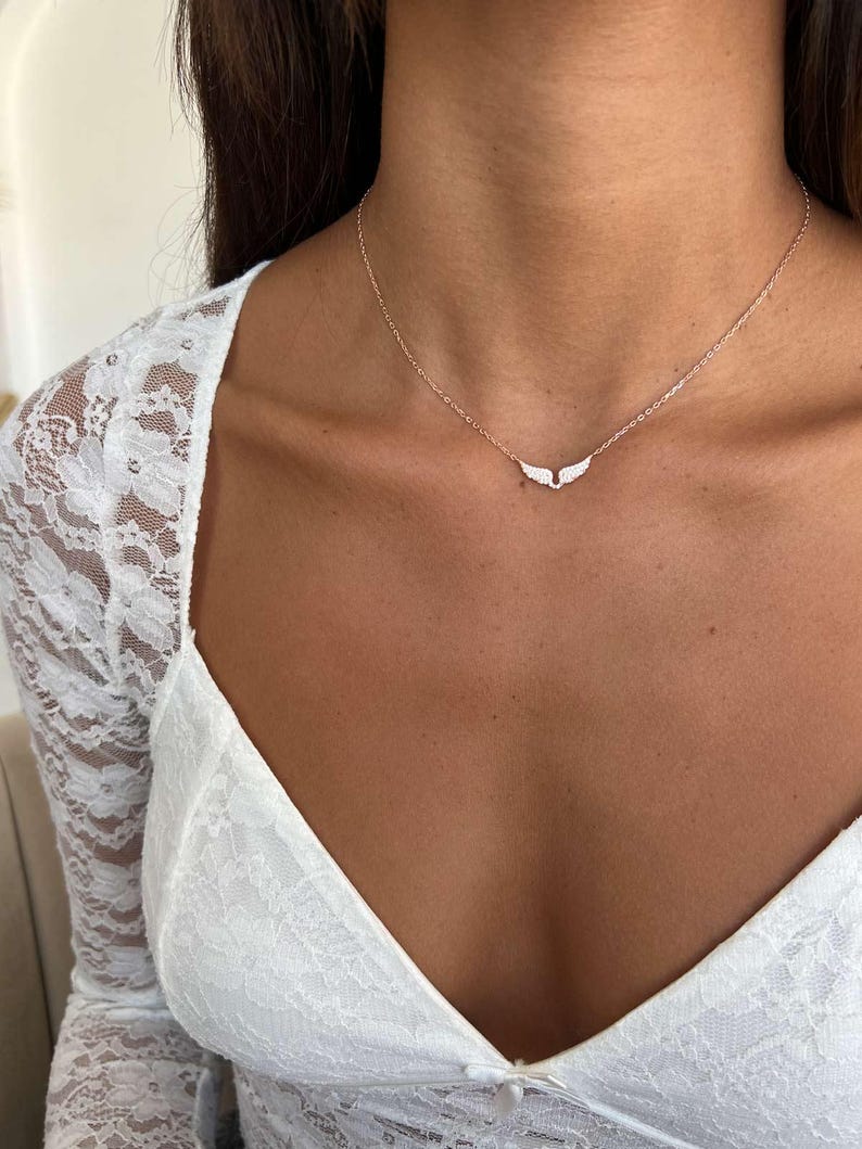 May include: A delicate necklace featuring a small, wing-shaped pendant. The pendant is adorned with small, sparkling stones. The necklace is gold-toned and rests on the neckline. The background includes a white lace top.