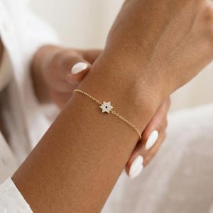 May include: A delicate gold bracelet featuring a star-shaped charm adorned with small, sparkling stones. The bracelet is on a slender chain, and the star has a dark center. The jewelry is worn on a wrist with manicured nails.