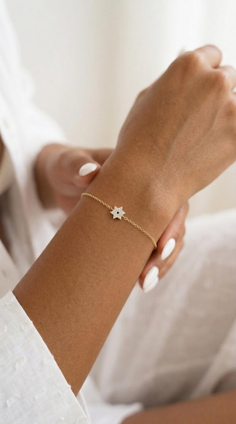 May include: A delicate gold bracelet featuring a star-shaped charm adorned with small, sparkling stones. The bracelet is on a slender chain, and the star has a dark center. The jewelry is worn on a wrist with manicured nails.