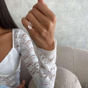 May include: A rose gold ring featuring a clear, pear-cut gemstone. The ring is worn on a finger, with a white lace sleeve in the background. The ring is a piece of jewellery.
