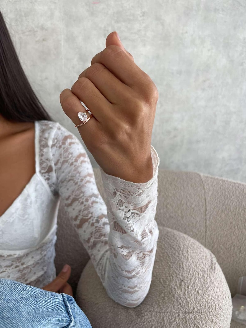 May include: A rose gold ring featuring a clear, pear-cut gemstone. The ring is worn on a finger, with a white lace sleeve in the background. The ring is a piece of jewellery.