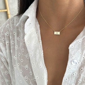 May include: A gold chain necklace with a small rectangular pendant featuring a delicate design. The necklace is worn by a person with long dark hair and a white blouse.